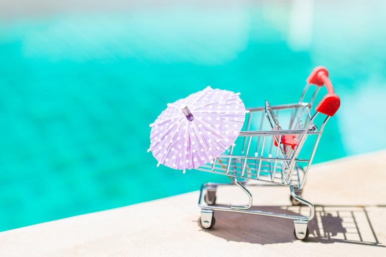 Leerer Einkaufswagen mit Papier-Regenschirm über unscharfen blauen Wasser (Pool) Hintergrund, Pool-Shopping-Konzept