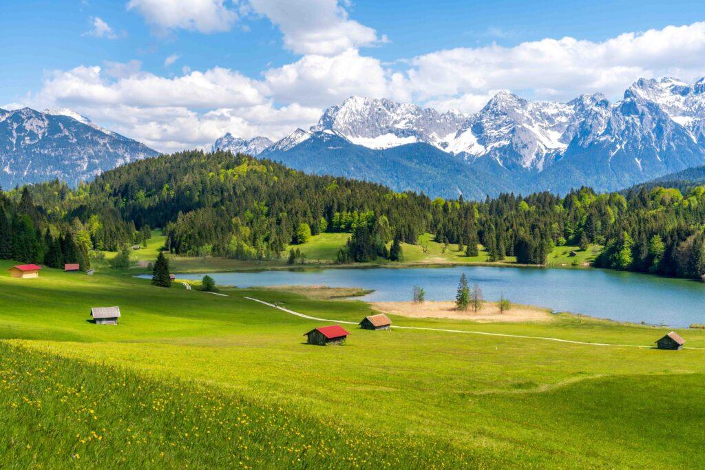 Der Geroldsee in Bayern, Deutschland, mit rustikalen Holzunterkünften und den majestätischen, schneebedeckten Karwendelbergen unter einem sonnigen blauen Sommerhimmel.